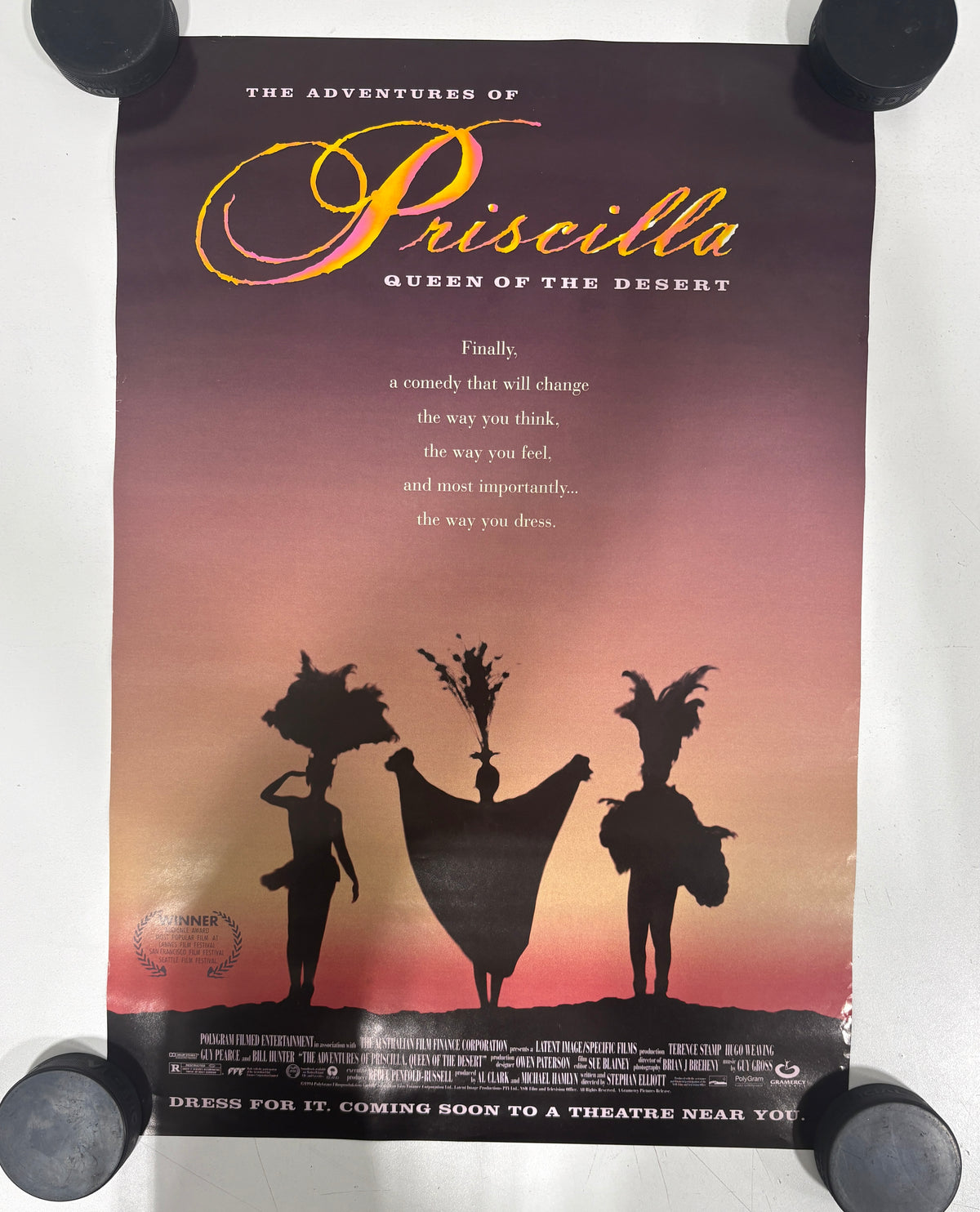 A vintage movie poster for 'The Adventures of Priscilla Queen of the Desert' displayed flat against a white background, held down at the corners by black weights.
