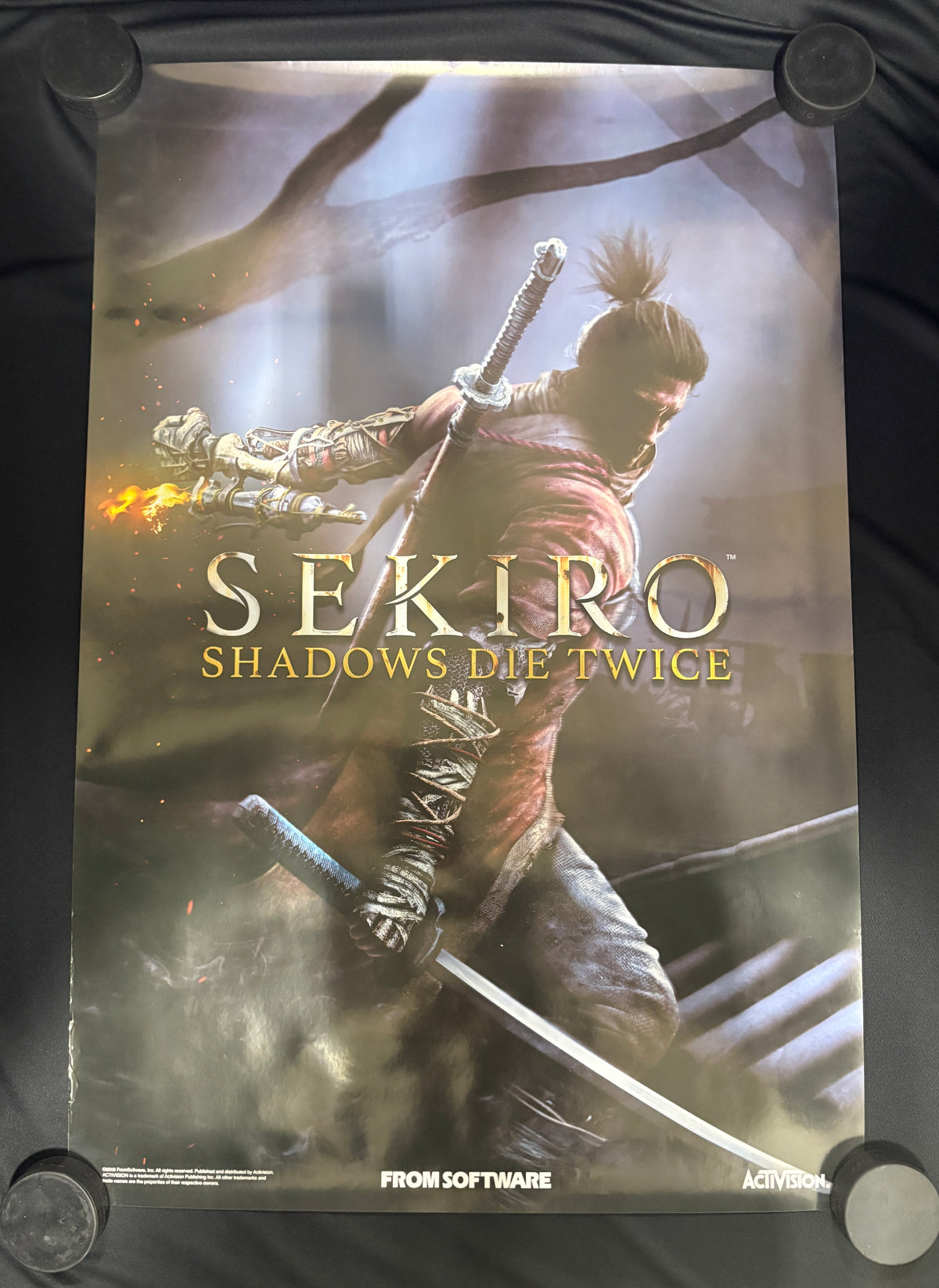 A printed Sekiro: Shadows Die Twice poster or art print displayed against a dark fabric background, with the samurai character and large title text visible.